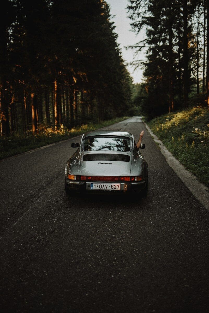 A silver sports car with a hand waving from the window on a forest road